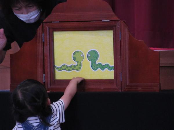 一人のお子さんが紙芝居の舞台の前で興味深く見ていて、蛇が2匹向き合っているところの紙芝居の写真
