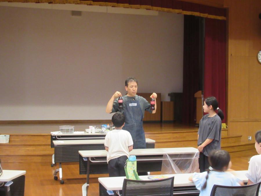 二人の児童が代表で出てきて、黒いペットボトルを振っているところ
